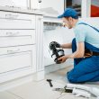 Male plumber in uniform installing disposer in the kitchen. Handywoman with toolbag repair sink, sanitary equipment service at home